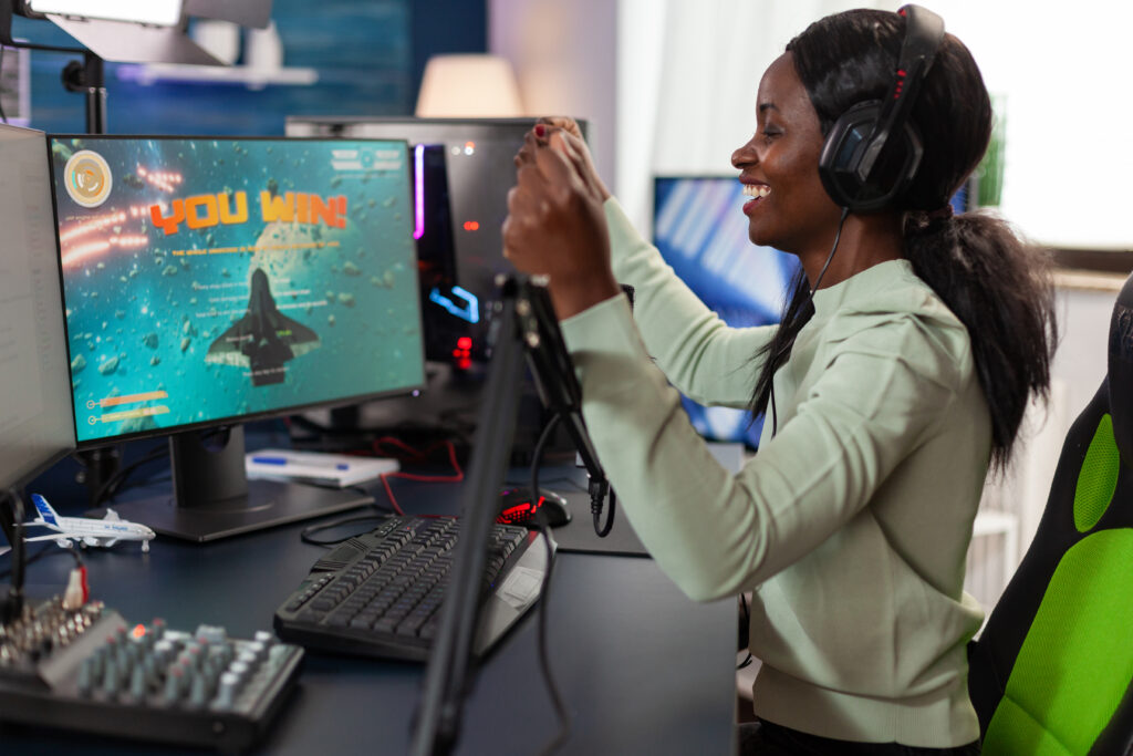 Happy African-American woman gamer wearing headset winning videogames competition talking into microphone with players. Excited plater playing space shooter games using RGB computer.